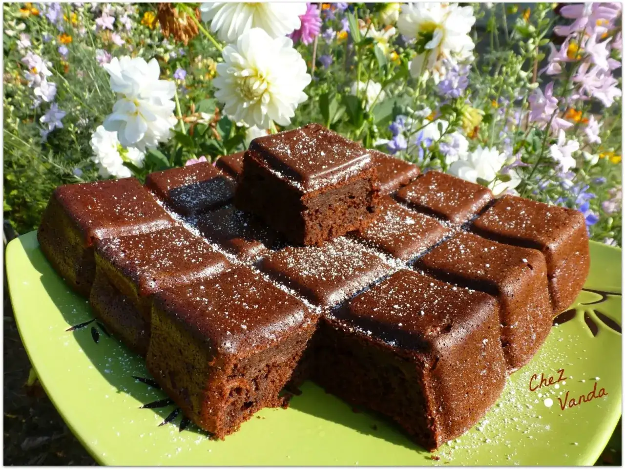 D&eacute;licieux g&acirc;teau courgette au chocolat, saupoudr&eacute; de sucre glace, sur une assiette verte devant un parterre de fleurs.