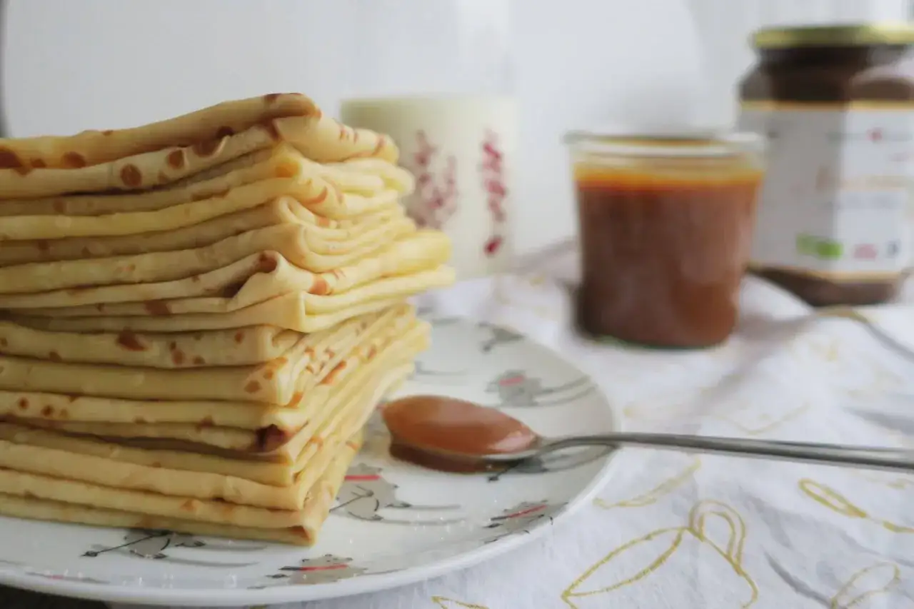 Une pile de cr&ecirc;pes moelleuses attend sur une assiette d&eacute;cor&eacute;e, avec une cuill&egrave;re de caramel et des pots en arri&egrave;re-plan.