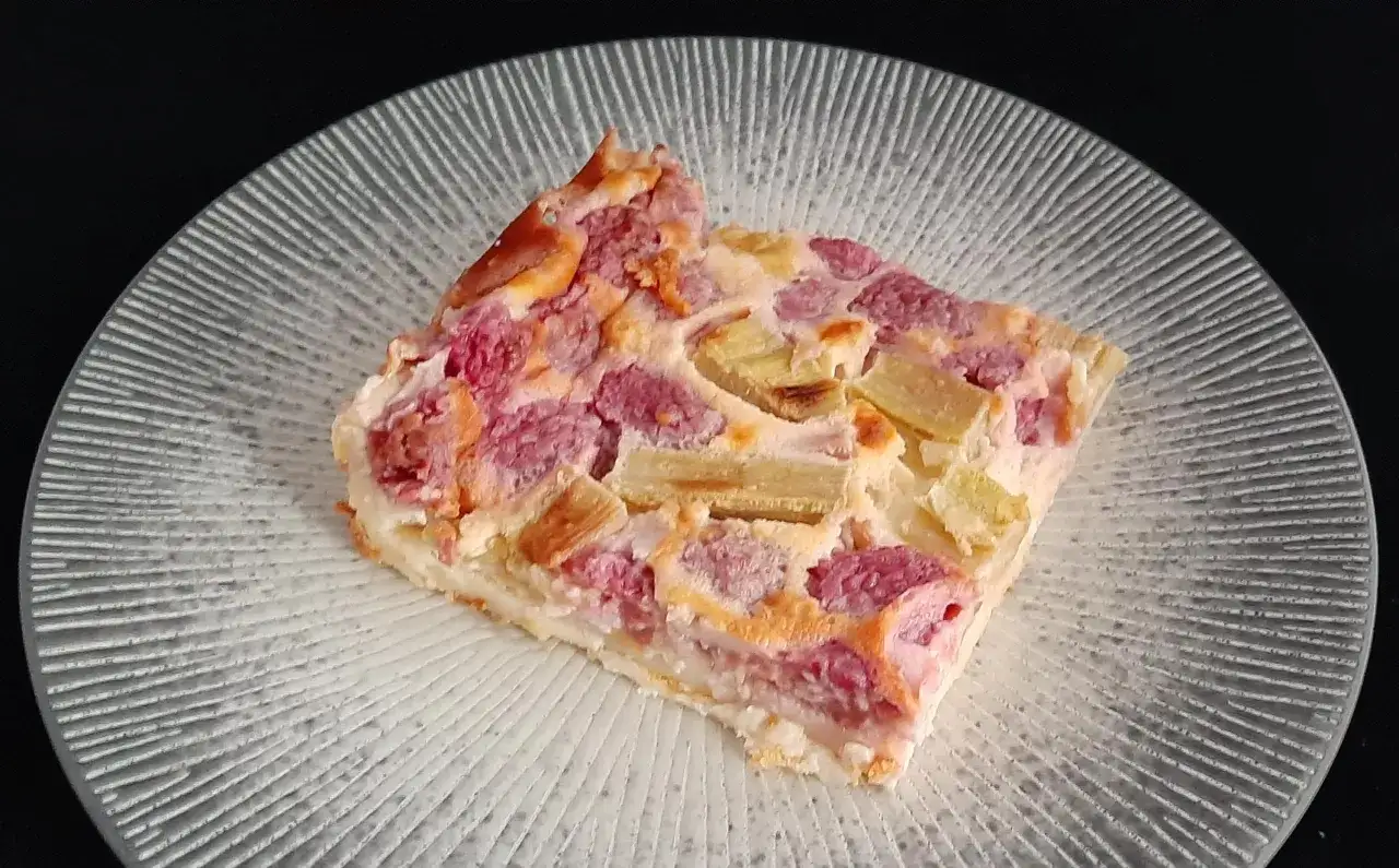 Une part de clafoutis rhubarbe aux framboises, dorée et appétissante, sur une assiette texturée.