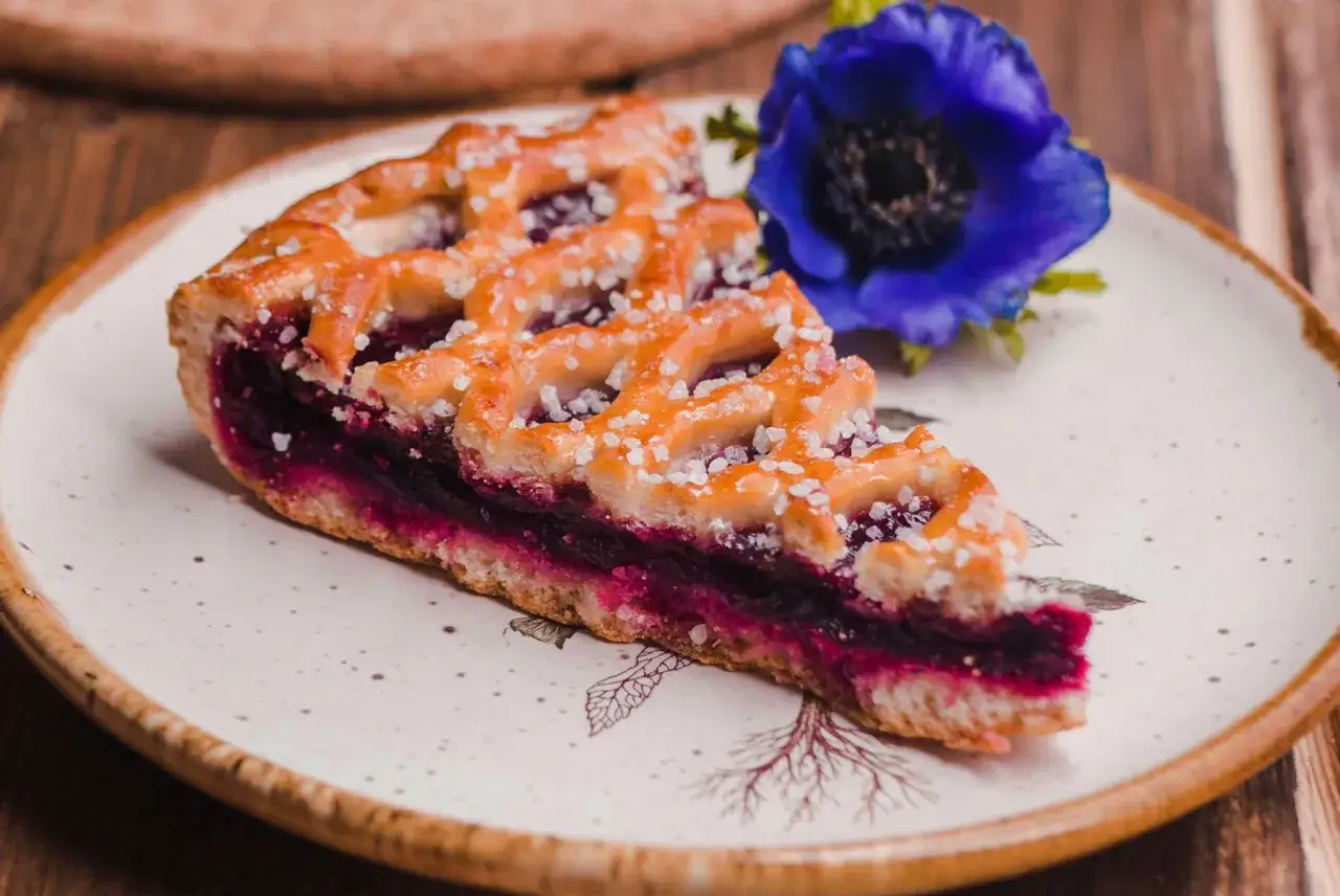 Une part de tarte aux cassis, garnie d'un croisillon doré et de gros cristaux de sucre, est servie sur une assiette décorée, à côté d'une fleur bleue.