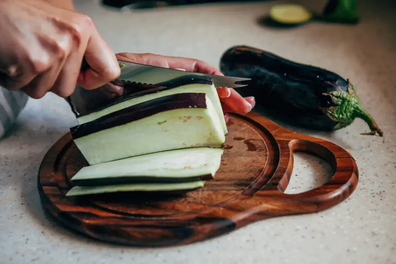 Une main coupe une aubergine en tranches sur une planche en bois. Préparation pour le temps de cuisson aubergine à l'eau.
