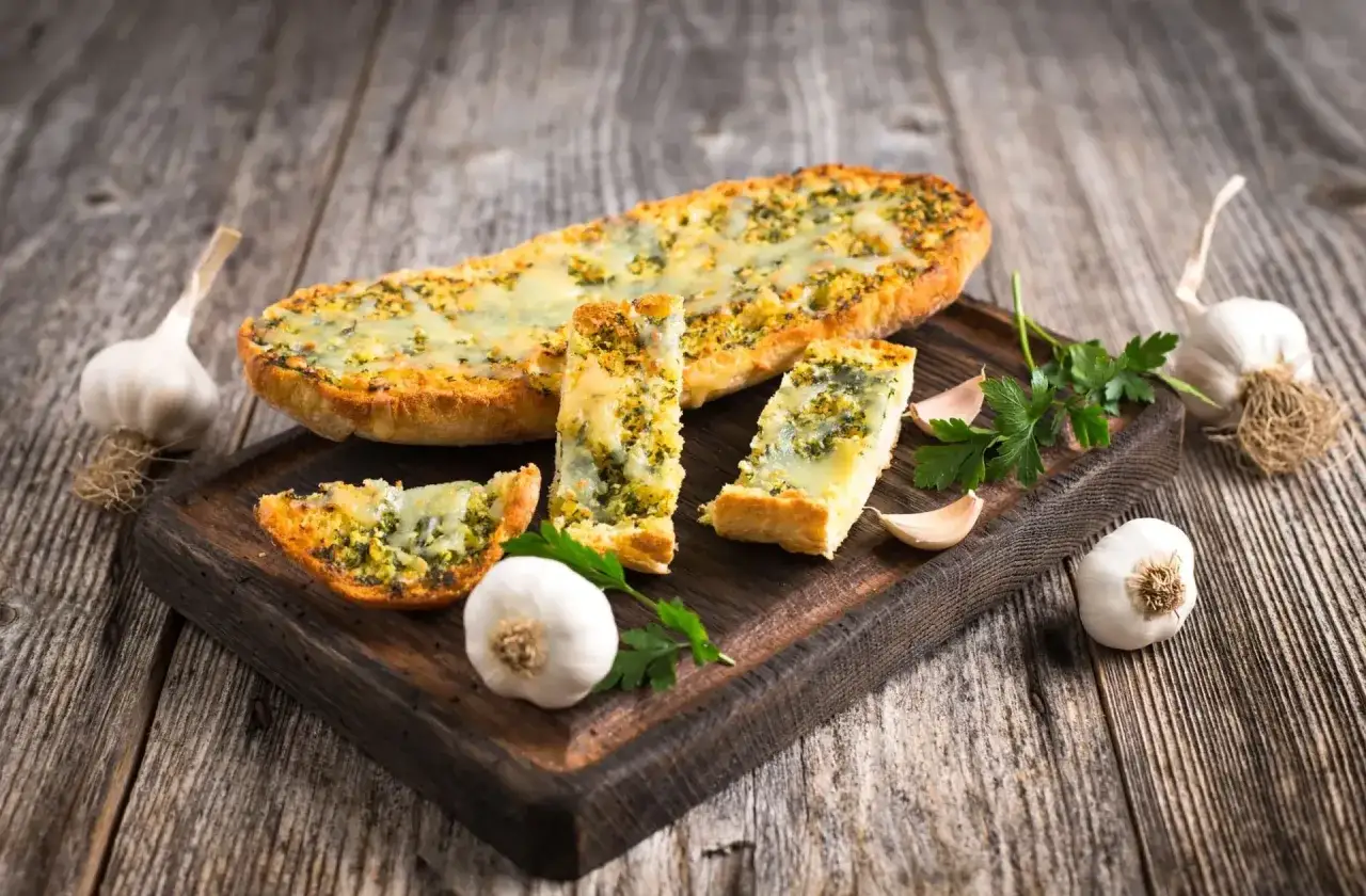 Tranches de pain &agrave; l'ail dor&eacute; et gratin&eacute;, garnies d'herbes et de fromage, pr&eacute;sent&eacute;es sur une planche en bois avec de l'ail frais et du persil.