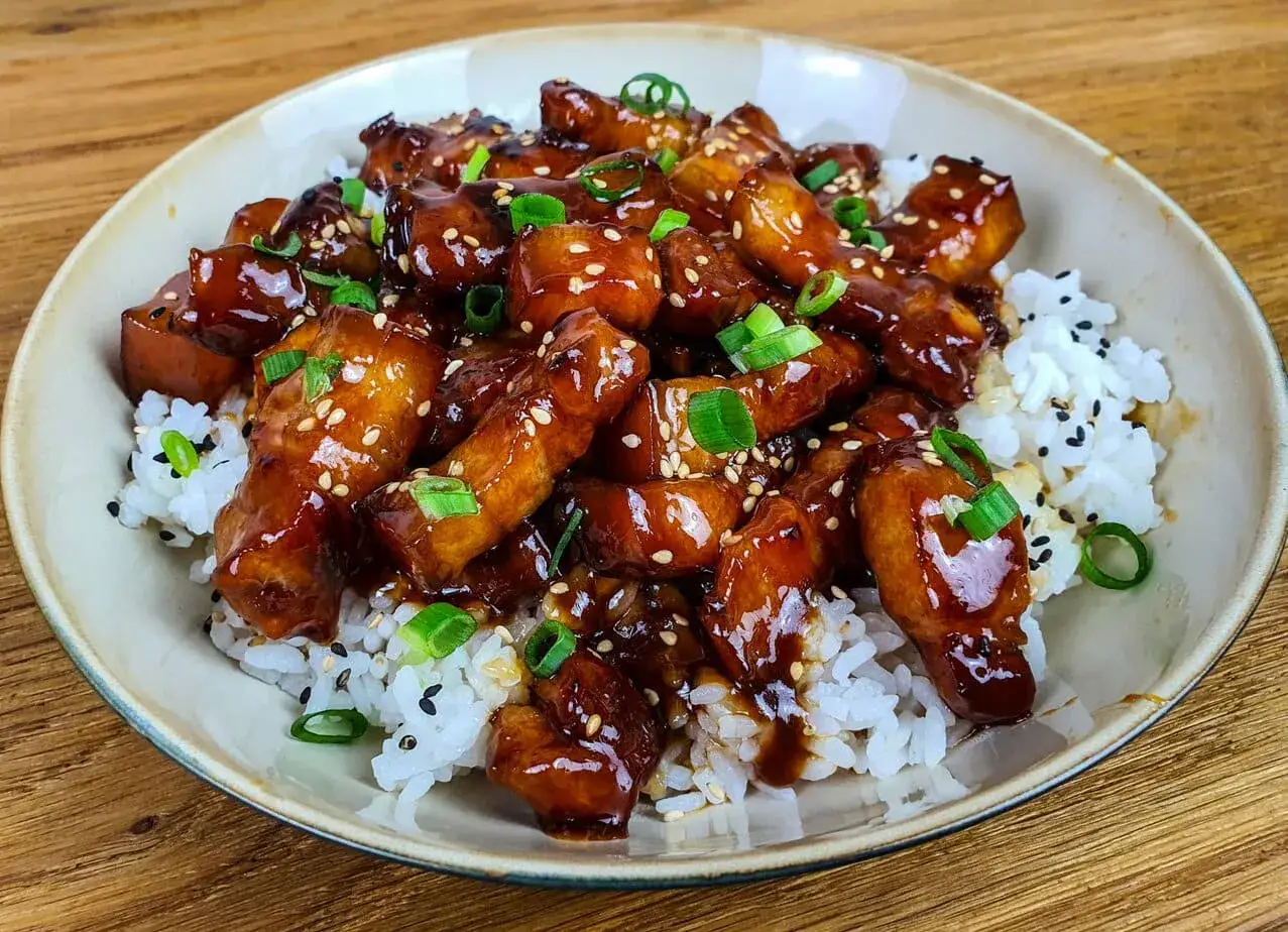 Un plat app&eacute;tissant de porc au caramel, garni de graines de s&eacute;same et d'oignons verts, servi sur un lit de riz blanc.