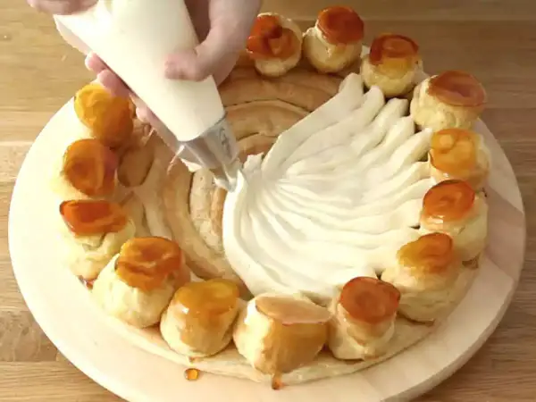 Préparation d'une recette crème chiboust Marmiton : on garnit un gâteau de crème à la poche à douille, entouré de petits choux caramélisés.