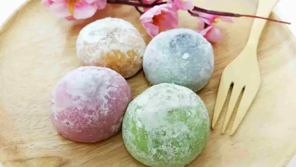 Quatre mochis colorés (rose, vert, bleu, jaune) saupoudrés de sucre glace, sur une assiette en bois avec une fleur rose. Une recette facile à réaliser !