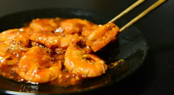 Gambas plancha enrobées d'une sauce orangée, une crevette est saisie par des baguettes.