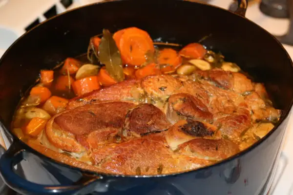 Rouelle de porc mijotant dans une cocotte avec des carottes, de l'ail et des herbes.