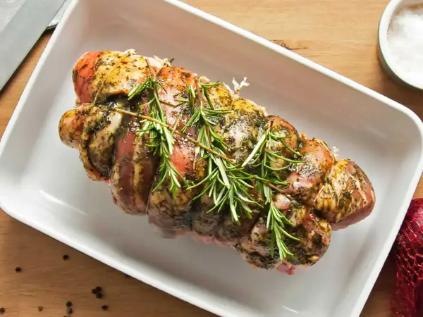 Un rôti de porc assaisonné et garni de romarin, prêt pour la cuisson au four dans un plat blanc.