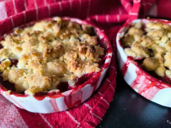 Deux petits ramequins de crumble aux quetsches, aux bords rouges et blancs, sur un torchon à rayures rouges.