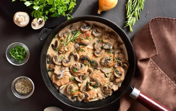 Une poêle remplie d'escalope à la crème et de champignons, garnie de persil frais et d'une branche de romarin.