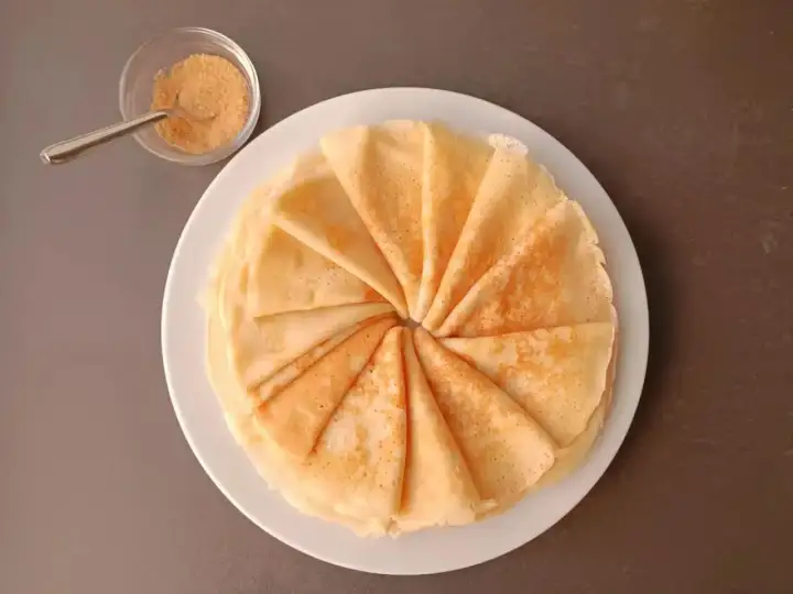 Une pile de crêpes moelleuses, pliées en éventail, sur une assiette blanche. À côté, un petit bol de sucre roux et une cuillère.